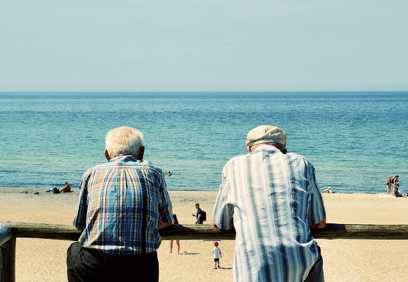 Zwei ältere Herren stehen an einem Zaun am Strand mit Blick aufs Mehr bei gutem Wetter.